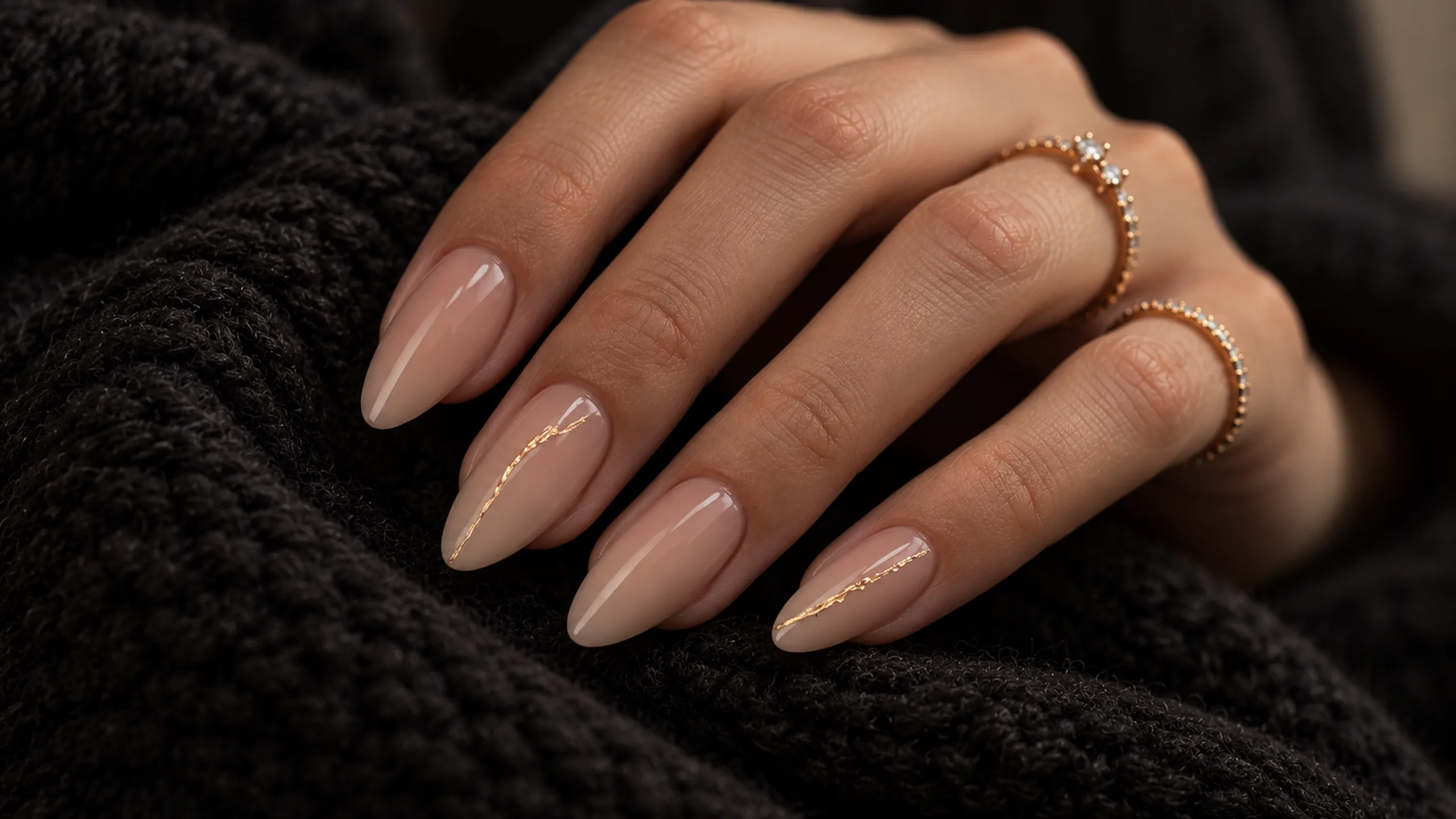 Nude-mauve almond nails with fine gold-foil lines and delicate gold rings on black knit fabric